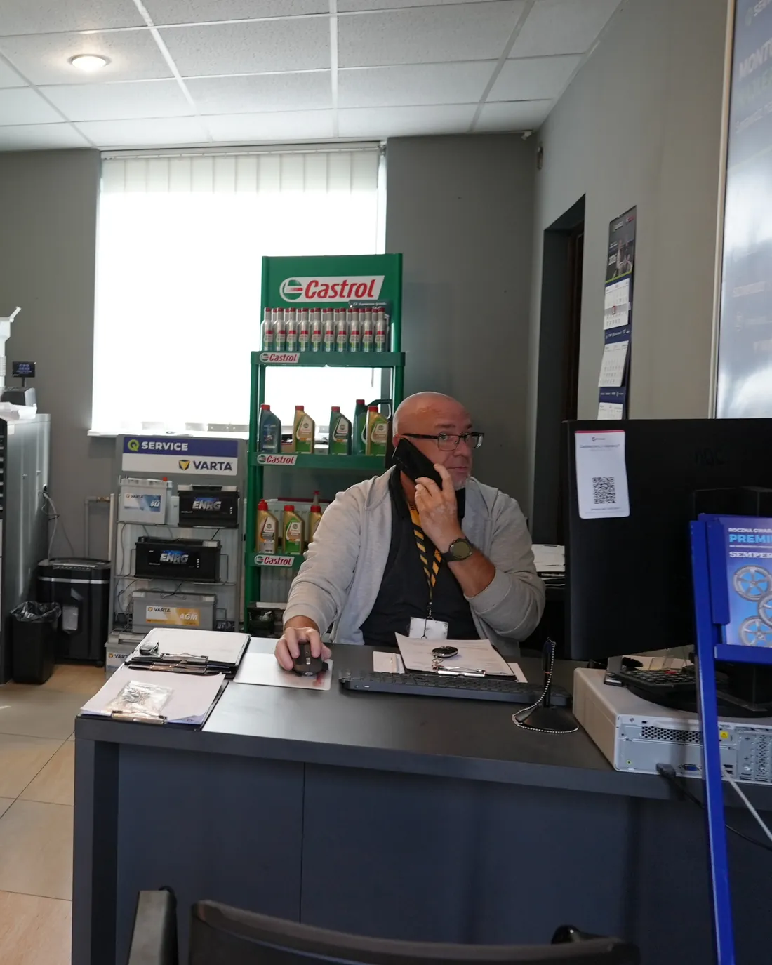 Biuro obsługi Bryksacar: pracownik przy biurku rozmawia przez telefon, za nim regał z olejami Castrol i akumulatorami