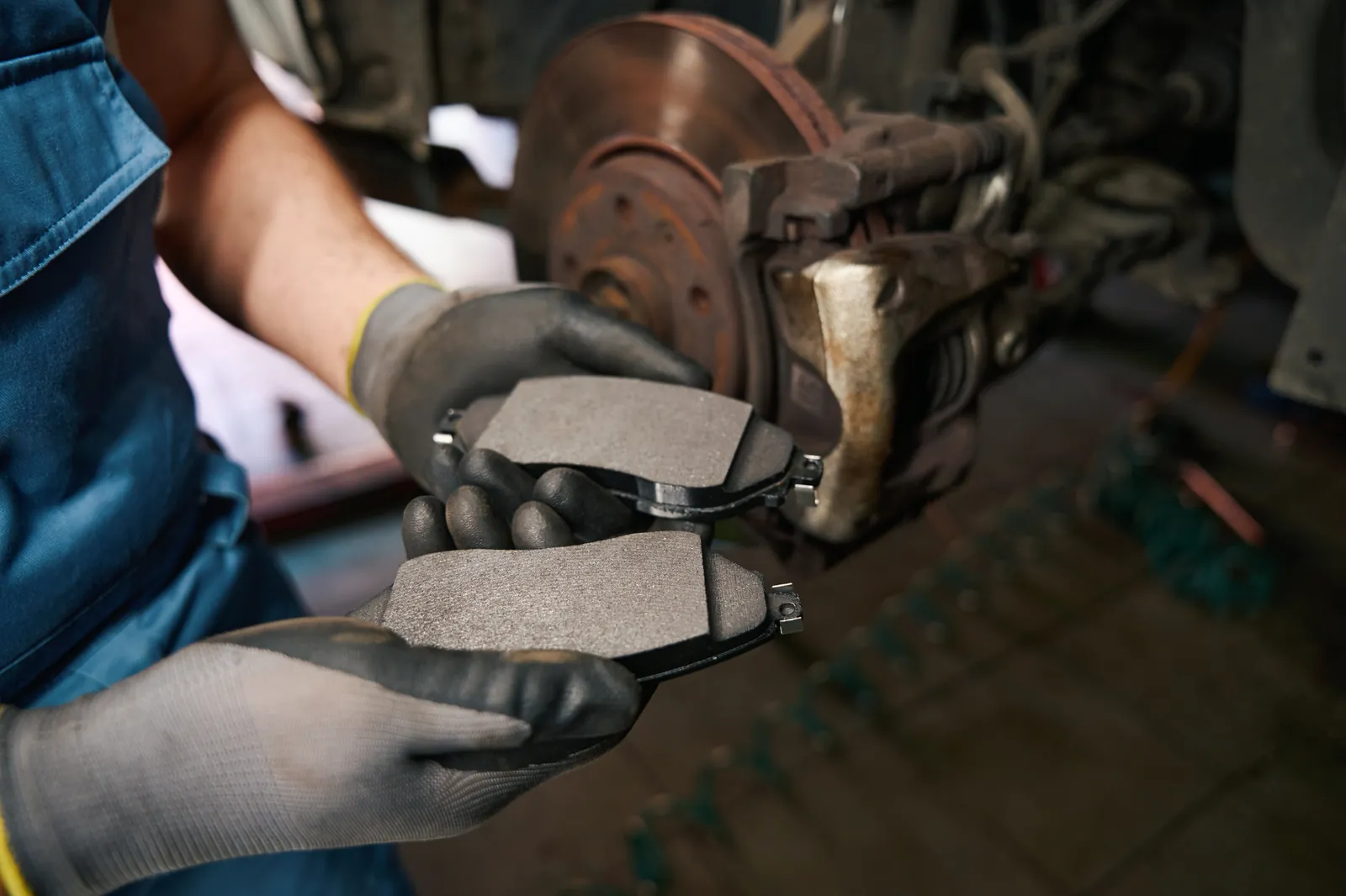 Mechanik w rękawicach trzyma dwa nowe klocki hamulcowe przy rozebranej tarczy